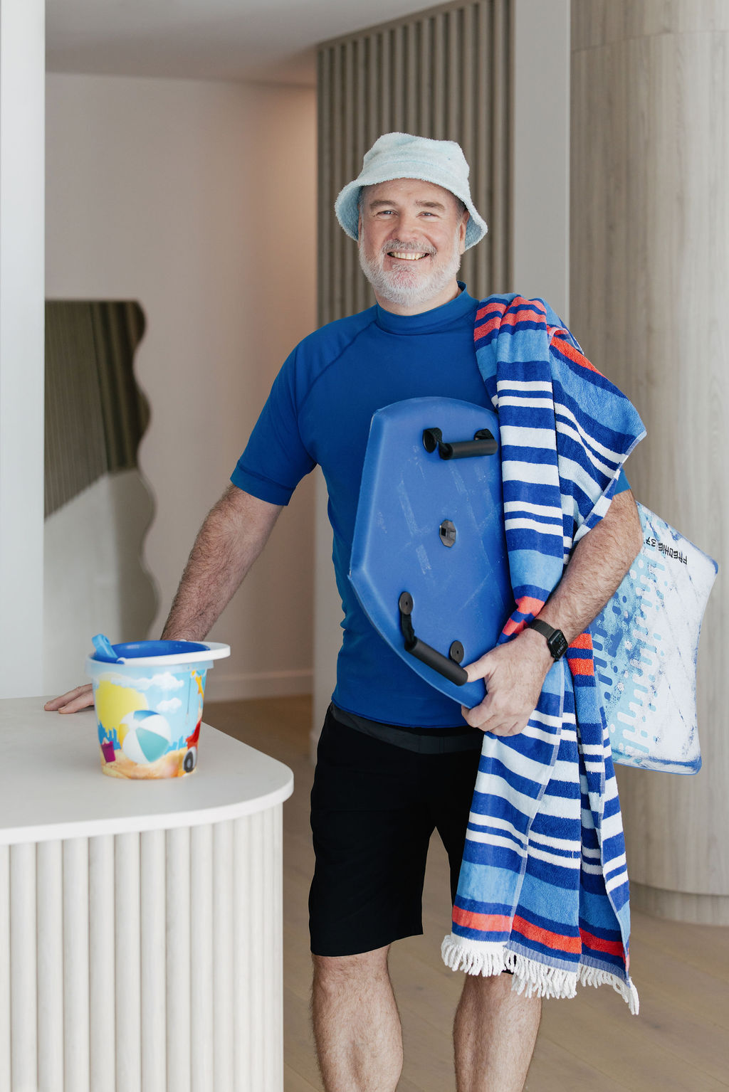 Australian in a blue hat, holding a body board and a stripy towel. Standing next to a bucket and spade.