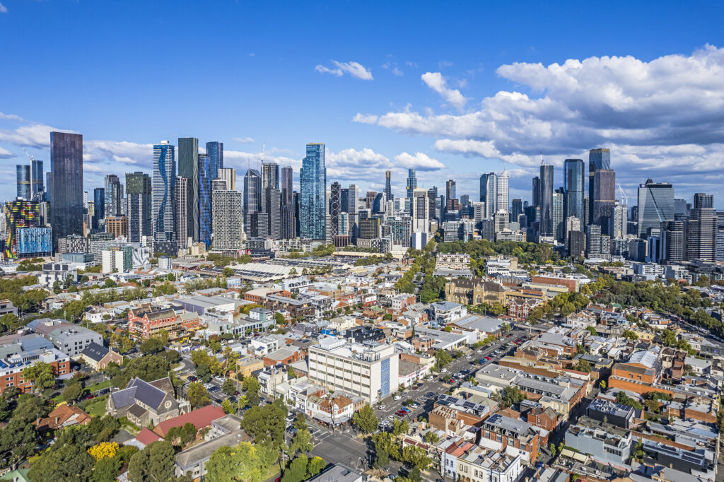 Aerial view of Melbourne's growing wide cluster of CBD skyscrapers taken from North Melbourne showing the contrast of old and new architecture in the foreground. Tree-lined streets low-rise buildings and parklands in established older suburb.