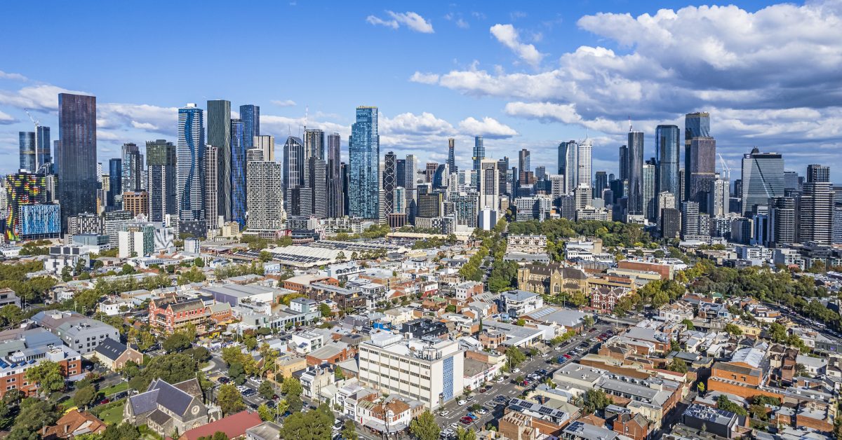 Aerial view of Melbourne's growing wide cluster of CBD skyscrapers taken from North Melbourne showing the contrast of old and new architecture in the foreground. Tree-lined streets low-rise buildings and parklands in established older suburb.